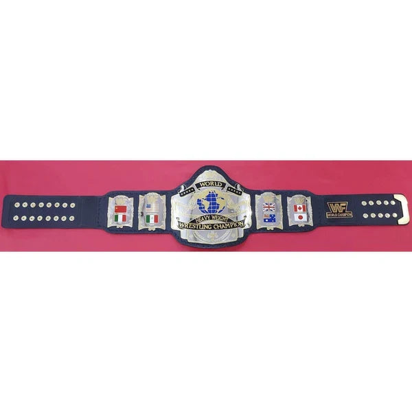 A wrestling championship belt displayed horizontally against a solid pink background. The belt features a navy blue strap with silver-toned metal plates, including a large central plate with a blue and gold graphic of a wrestler inside a ring, and smaller plates with various country flags.