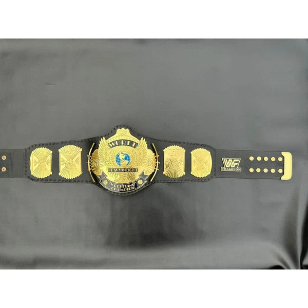 A black and gold WWE Championship belt laid flat on a dark gray fabric background, featuring the central gold-plated WWE Championship medallion and matching side plates.