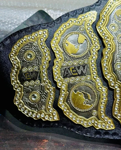 Close-up of a black wrestling championship belt with ornate gold and silver AEW plates, detailed with small clear rhinestones and raised world map emblems.