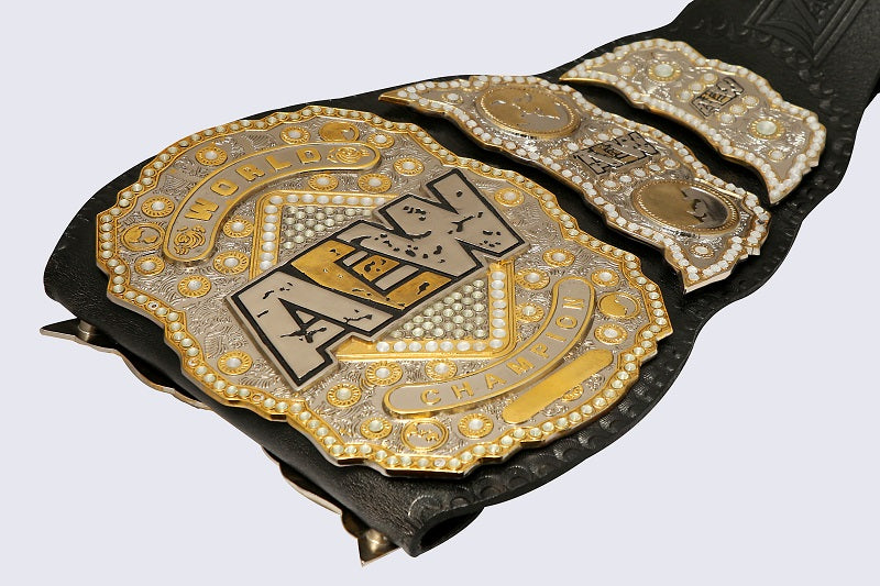 A close-up of a black leather championship belt with ornate gold and silver metal plates, featuring the AEW (All In The Night) logo on the main plate, set against a light gray background.