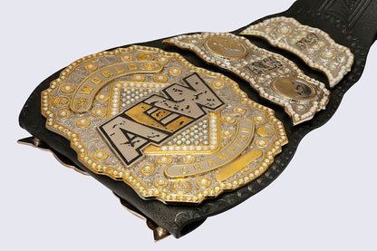 A close-up of a black leather championship belt with ornate gold and silver metal plates, featuring the AEW (All In The Night) logo on the main plate, set against a light gray background.