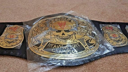 A WWE Heavyweight Championship belt with a large gold skull centerpiece and two smaller matching side plates, laid on a black strap over a tan carpet. The center section is wrapped in clear plastic.