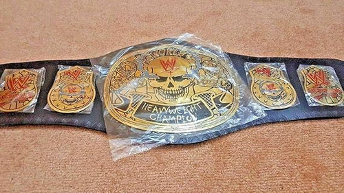 A WWE Heavyweight Champion belt with gold-toned metal plates and a black strap, shown wrapped in clear plastic on a tan carpeted floor. The center plate features a skull and the WWE logo, with smaller matching plates on both sides.