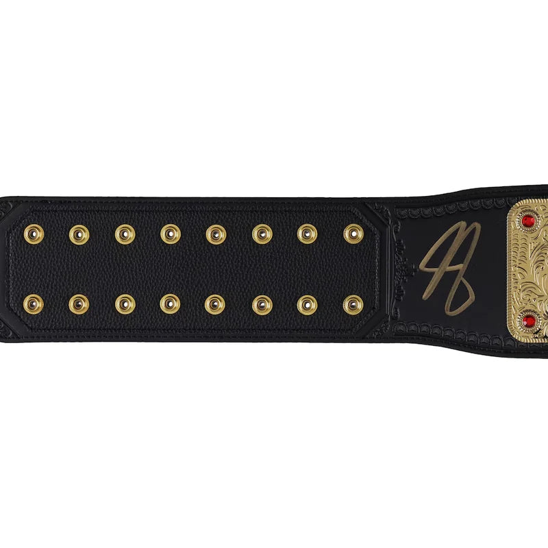A close-up of a black leather championship belt strap with gold-tone studs, a gold signature, and a gold-toned metal plate with red gem accents, shown against a plain white background.