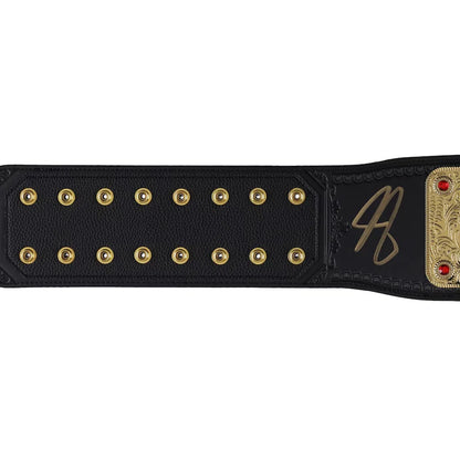 A close-up of a black leather championship belt strap with gold-tone studs, a gold signature, and a gold-toned metal plate with red gem accents, shown against a plain white background.