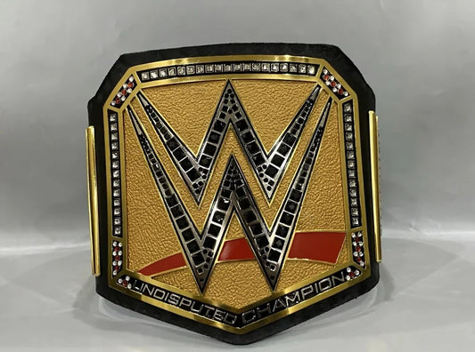 A WWE Undisputed Champion belt featuring a textured gold center with a large black and silver 'W' logo, bordered by rhinestones and red accents, set against a light gray background.