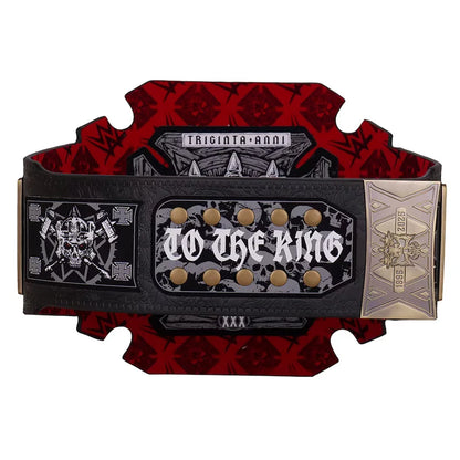 A championship-style belt buckle plaque with the words 'TO THE KING' in gothic lettering, surrounded by gold studs. The plaque is mounted on a black strap with red and black patterned backing, and a gold-toned end piece reads 'TRIGINTA ANNI'.