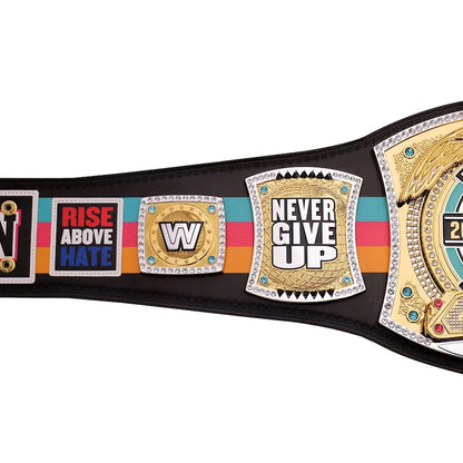 A close-up of a championship belt with multiple decorative plates. The belt is black with a colorful stripe pattern in teal, pink, and orange, featuring gold and silver plates with text including 'RISE ABOVE HATE', 'NEVER GIVE UP', and a large gold winged plate on the right.