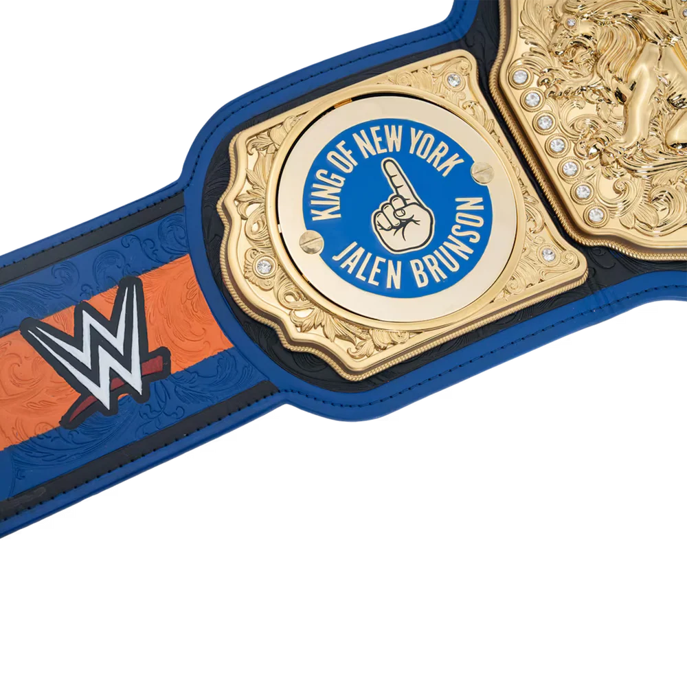 Close-up of a WWE championship belt featuring a gold central plate with a blue circle reading 'King of New York Jalen Brunson' and a raised middle finger hand icon, with blue and orange strap panels and a WWE logo on a white background.