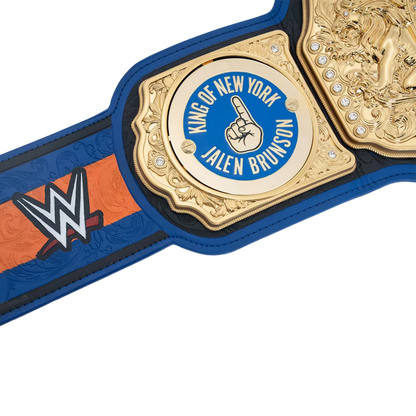Close-up of a WWE championship belt featuring a gold central plate with a blue circle reading 'King of New York Jalen Brunson' and a raised middle finger hand icon, with blue and orange strap panels and a WWE logo on a white background.