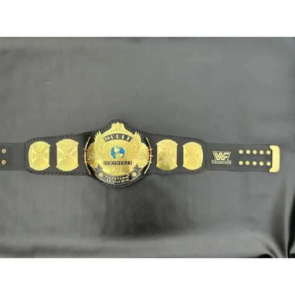 A black and gold WWE Championship belt laid flat on a dark gray fabric background, featuring the central gold-plated WWE Championship medallion and matching side plates.