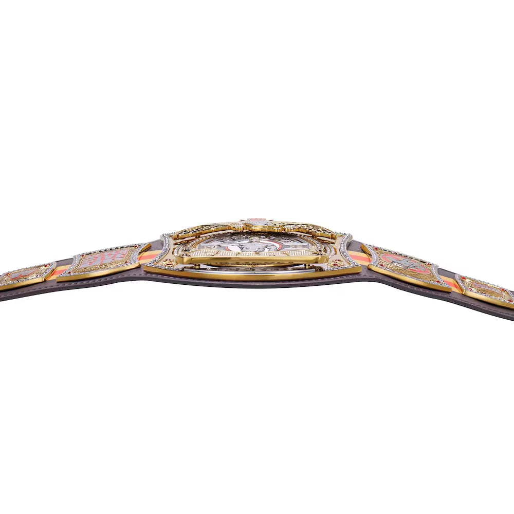 A close-up of a championship-style belt across a white background, showing ornate gold-toned metal plates, a central buckle area with a clear stone, and a dark strap extending off both sides.