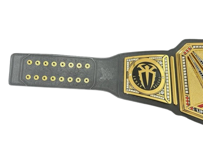 A close-up of a championship belt strap with a black leather-like base and ornate gold-tone metal plates, shown on a plain white background.