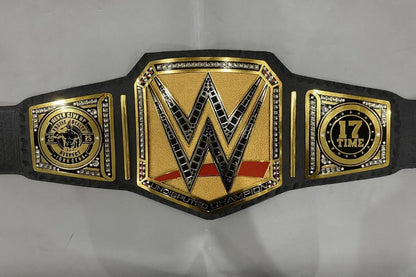 A WWE championship belt with a black strap and gold-toned metal plates, featuring the large central WWE 'W' logo, a 17-time championship emblem on the right, and a smaller emblem on the left, photographed against a light gray background.