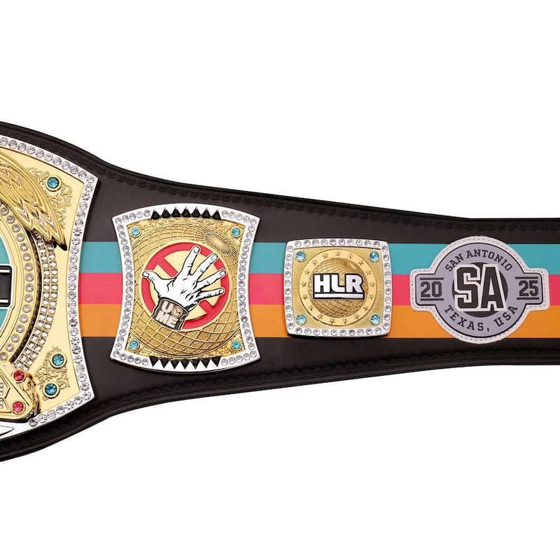 A close-up of a championship belt with multiple ornate metal plates, including a gold plate with a hand graphic, a gold HLR plate, and a San Antonio 2025 SA plate, set against a black strap with turquoise, pink, and orange stripes.