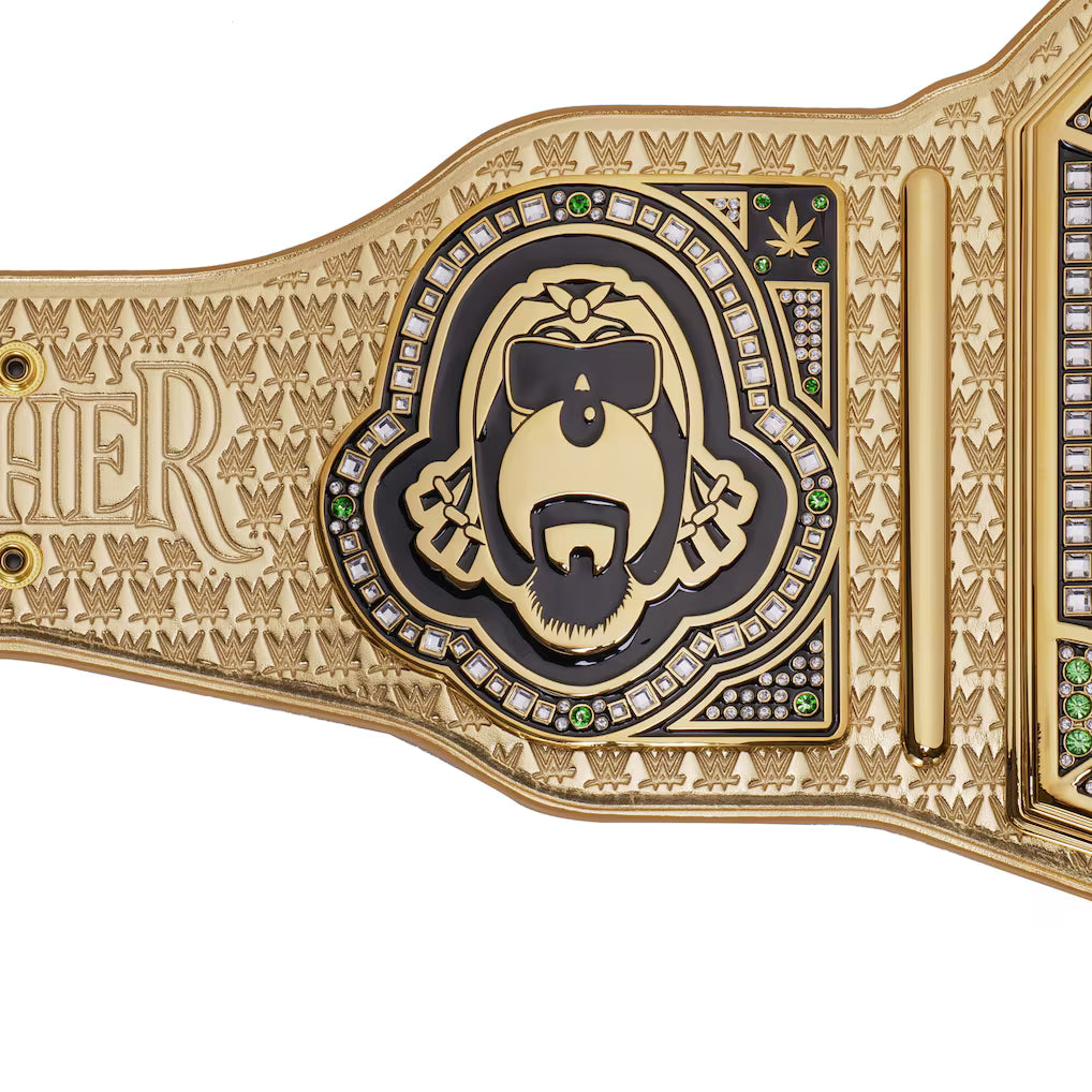 A close-up of a gold and black metal championship-style belt plate with an engraved gorilla face wearing sunglasses, surrounded by small clear stones and green gem accents, on a white background.