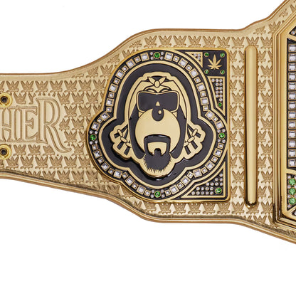 A close-up of a gold and black metal championship-style belt plate with an engraved gorilla face wearing sunglasses, surrounded by small clear stones and green gem accents, on a white background.
